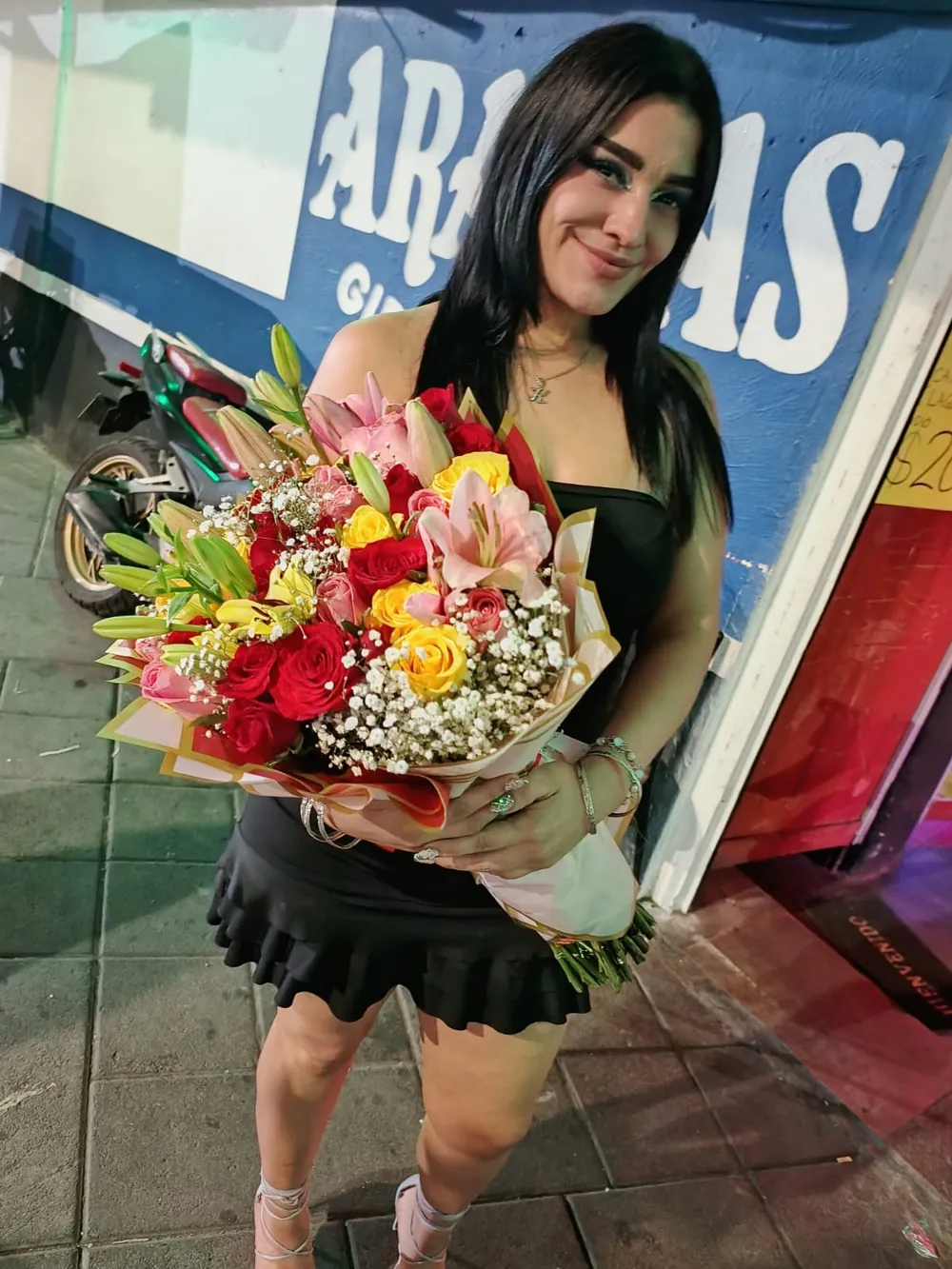 Joven sonriendo con un ramo de lilis, rosas y girasoles en la calle