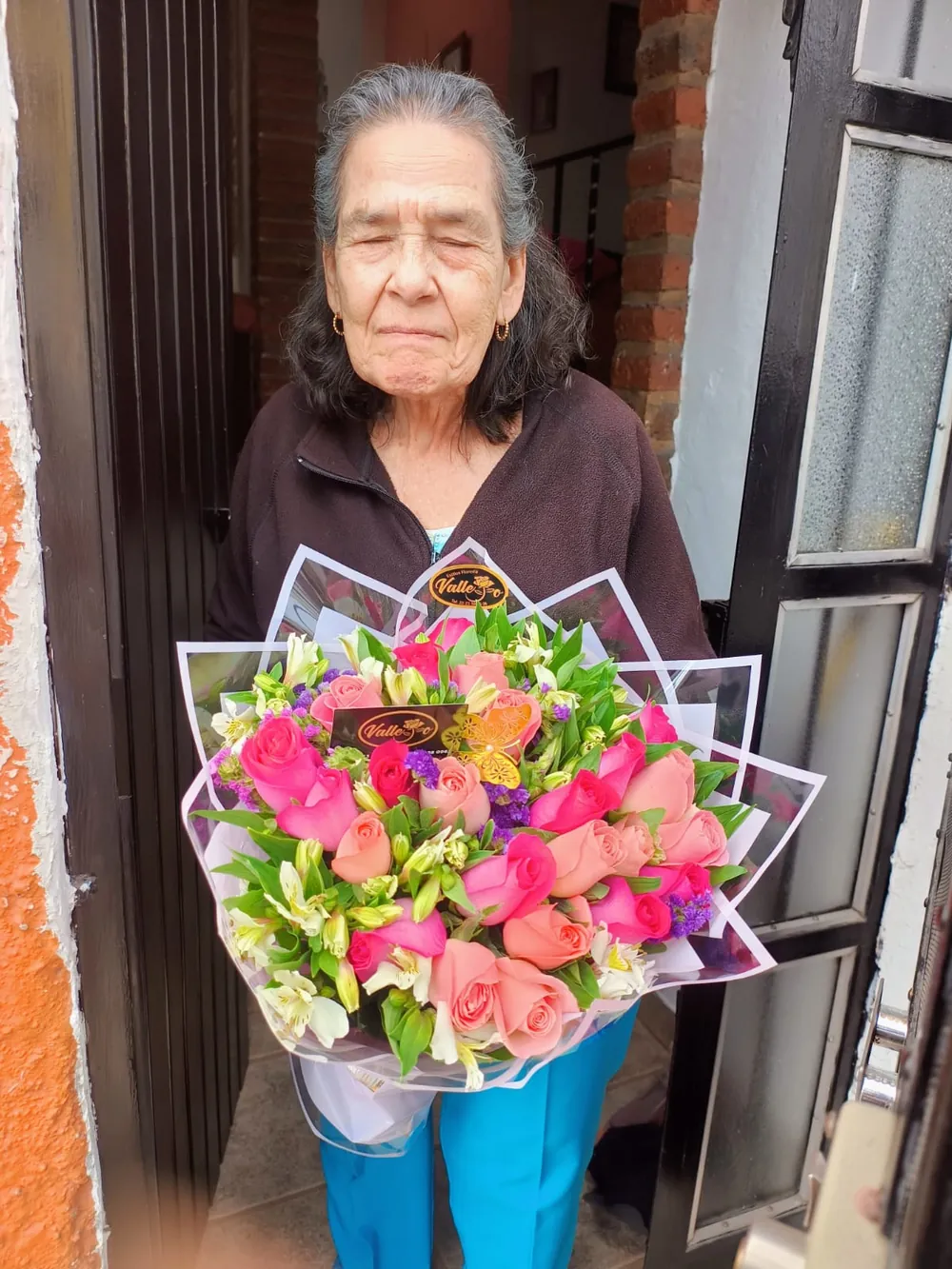 Señora sonriendo con un ramo de rosas y flores de colores en la puerta de su casa
